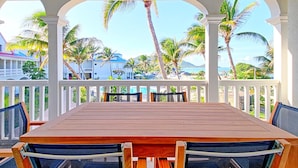 A covered outdoor dining area with a wooden table and chairs, white railings, and a view of palm trees and buildings.