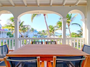 A covered outdoor seating area with a wooden table and chairs, overlooking a beach with palm trees and a clear sky.