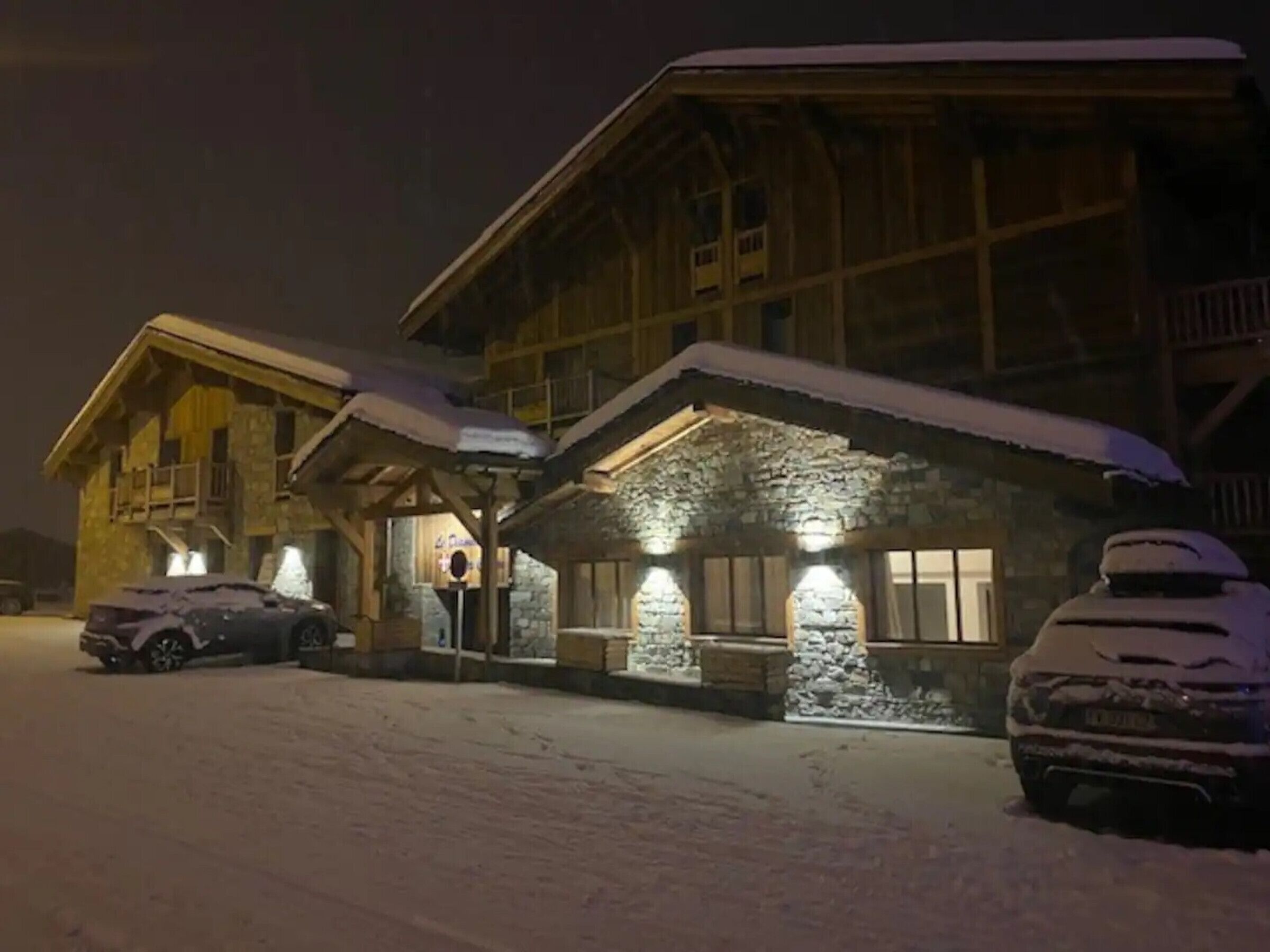 Un edificio in legno ricoperto di neve con facciata in pietra, finestre illuminate e auto parcheggiate.