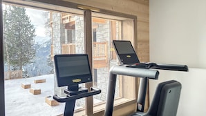 Ein Fitnessraum mit Laufbändern und einem Ergometer, großen Fenstern und Blick auf einen verschneiten Berg.