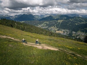 Trilha de mountain bike com dois ciclistas, cercada por vegetação exuberante e uma cidade no vale abaixo.