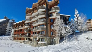 Ein mehrstöckiges Hotel mit Balkonen und einer Steinfassade, umgeben von schneebedeckten Bäumen und blauem Himmel.
