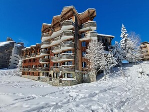 A multi-story hotel with balconies and a stone facade, surrounded by snow-covered trees and a clear blue sky.