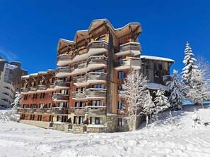 A multi-story wooden building with balconies, surrounded by snow-covered trees and a clear blue sky.