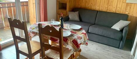 A dining area with a wooden table set for two, a grey sofa, and a wooden cabinet against a wooden wall.