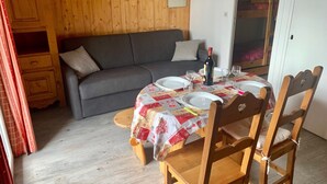 Une pièce chaleureuse avec un plancher en bois, un canapé gris, une table à manger dressée avec des assiettes et des verres, et une bouteille de vin.