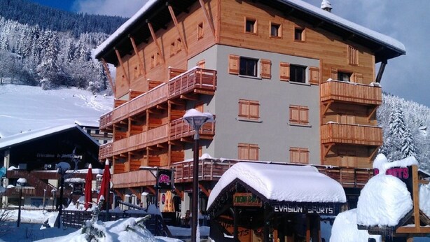 Un bâtiment en bois à plusieurs étages, doté de balcons, qui ressemble probablement à un hôtel ou à un complexe touristique, entouré d’arbres couverts de neige et surplombant un ciel dégagé.
