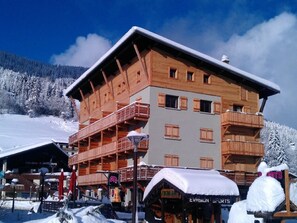 A multi-story wooden building with balconies, likely a hotel or resort, surrounded by snow-covered trees and a clear sky. - Cozy Studio in Les Gets, Parking Included, 4 People (Les Gets)