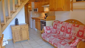 A compact kitchen with wooden cabinets, a microwave, and a stove. A wooden staircase leads to another level. A red and white patterned sofa is in the living area. - Cozy apartment in Morillon, 6 people, near Criou (Morillon Village)