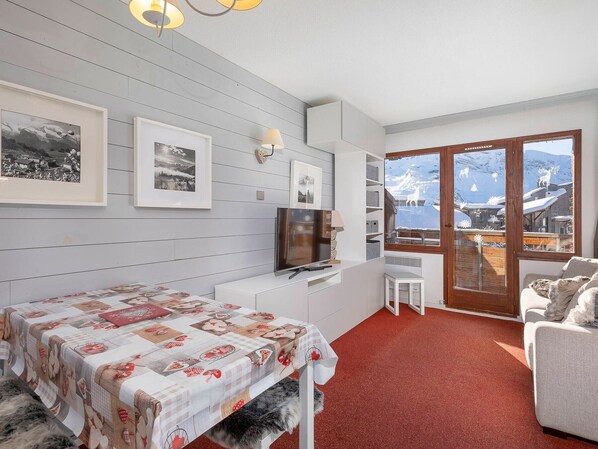 A cozy room with a bed, a TV, a desk, and a chair. There are framed pictures on the wall and a window with a view of a snowy landscape.