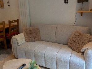Ein beiges Sofa mit zwei flauschigen Kissen, ein Holzstuhl, ein kleiner Tisch mit Fernbedienung und eine Pflanze.