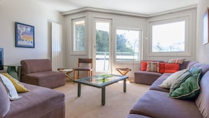 A living room with two sofas, a coffee table, and a chair. There are large windows and a door leading outside.
