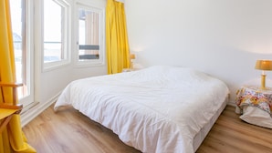 A bedroom with a large bed, a window with yellow curtains, a bedside table with a lamp, and a small table with a colorful tablecloth.