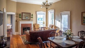 A spacious living room with a fireplace, a dining area with a table set for four, and a large chandelier. - The Mildred Haus (Mason)
