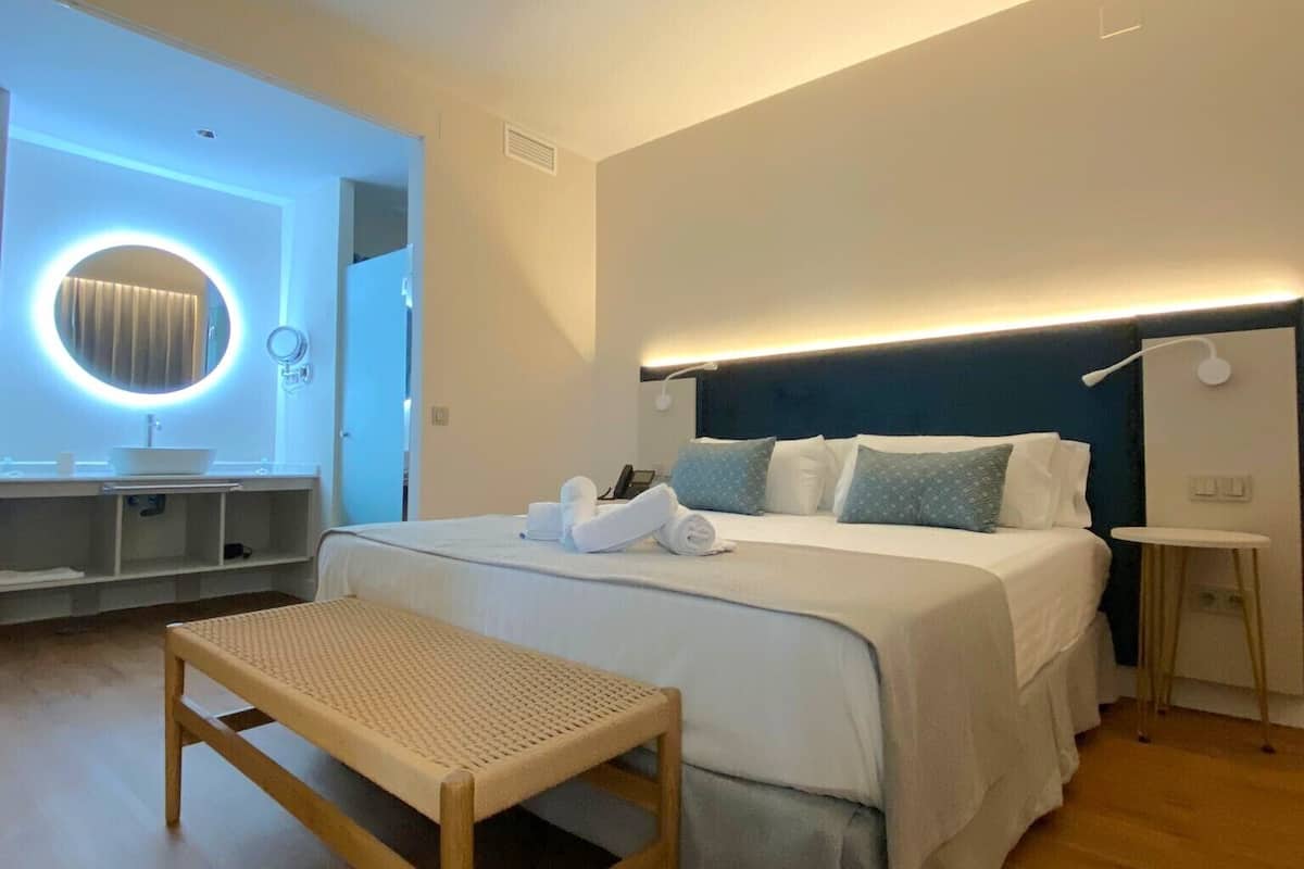 Una habitación de hotel moderna con una cama grande, mesita de noche y un espejo redondo con iluminación.