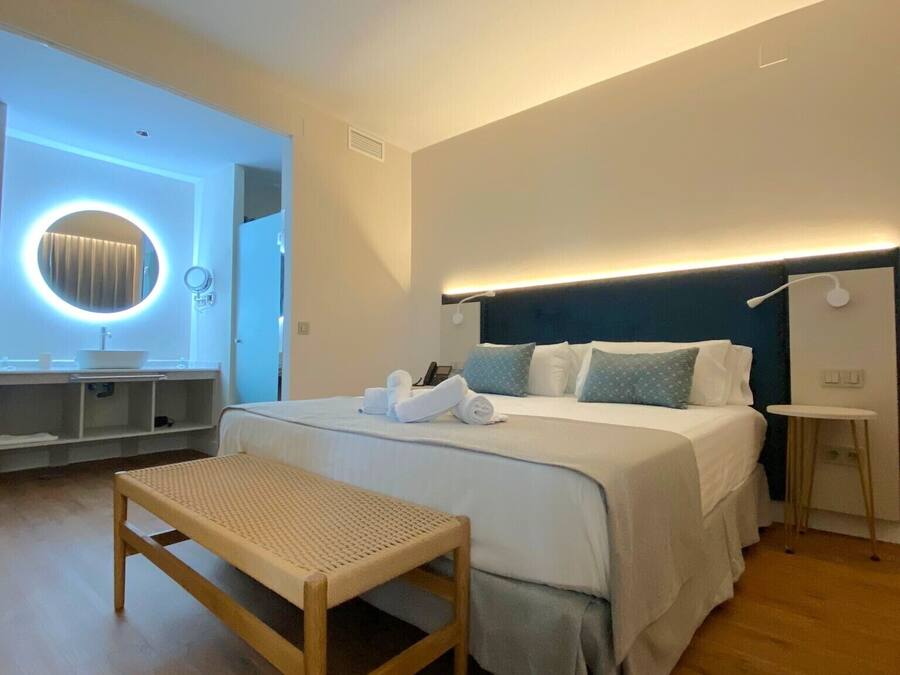 Una habitación de hotel moderna con una cama grande, mesita de noche y un espejo redondo con iluminación.