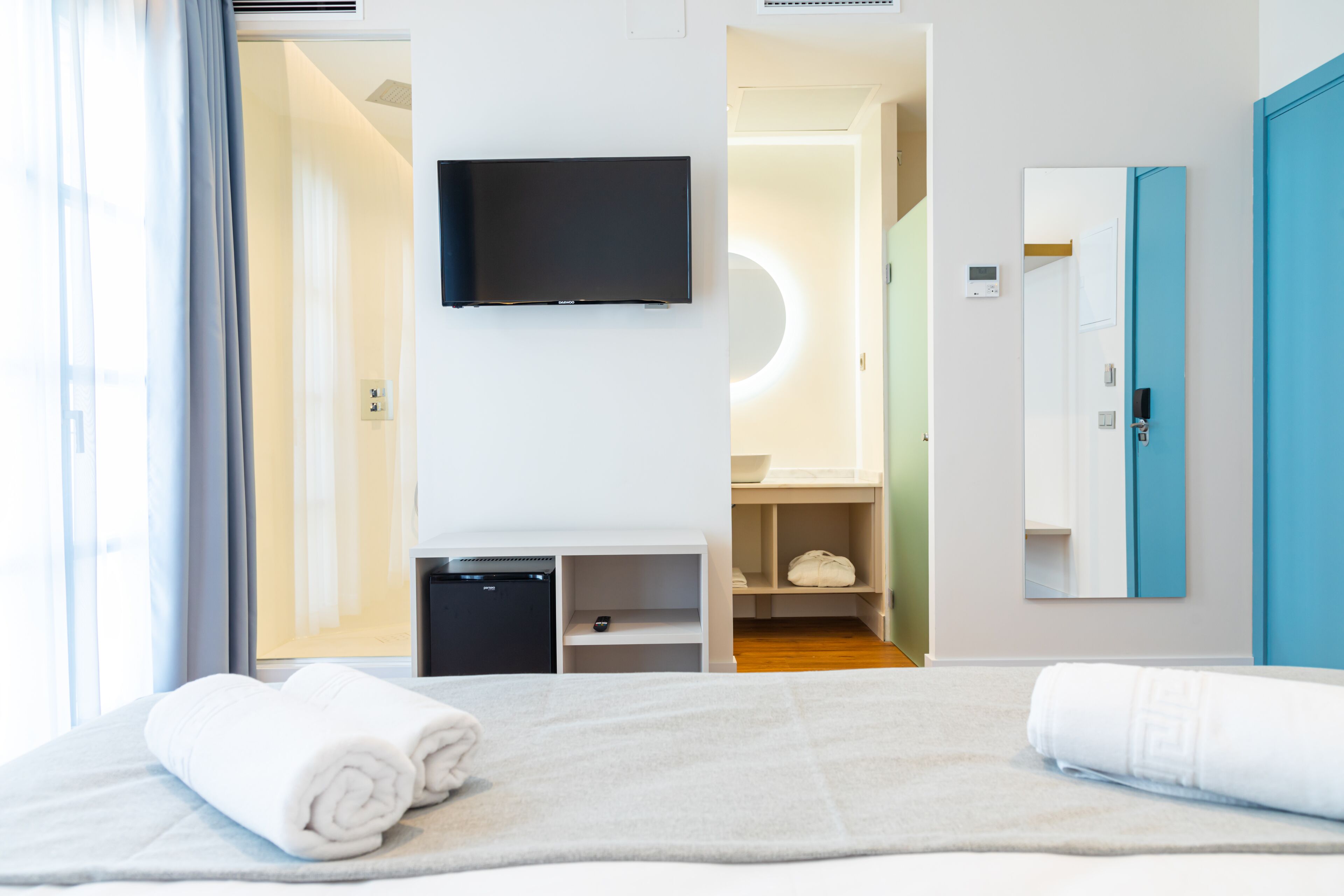 A modern hotel room with a bed, a flat-screen TV, a walk-in closet, and a bathroom with a mirror and towel rack.