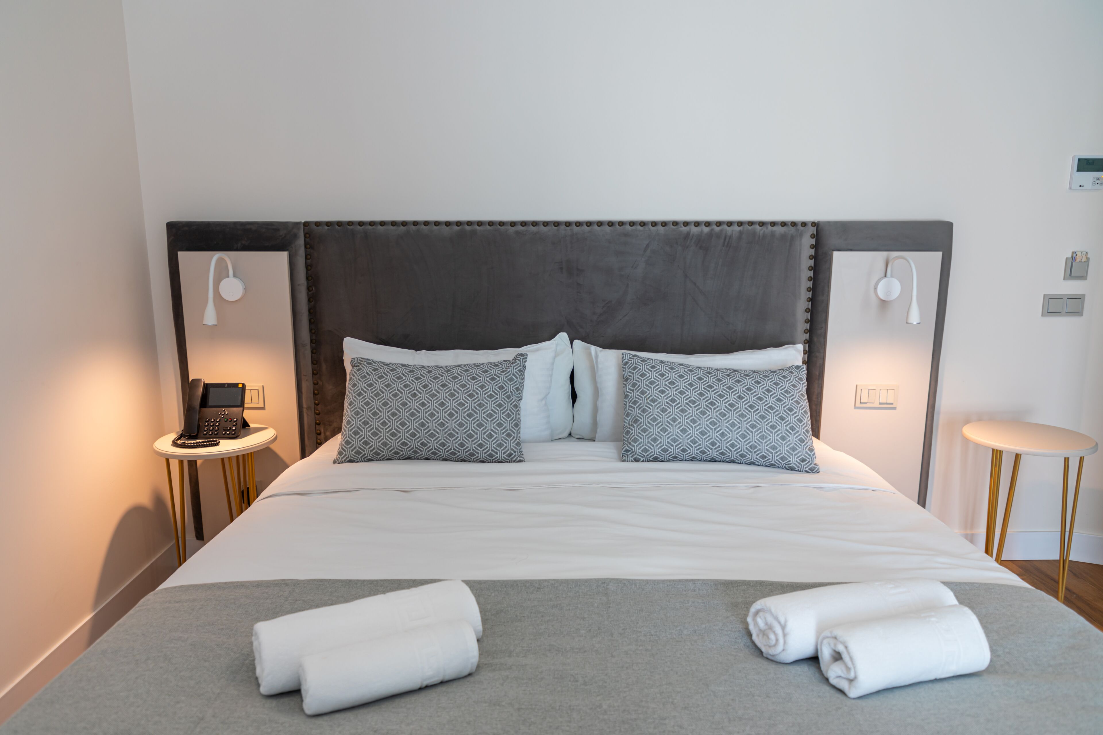 Una cama bien hecha con cabecera gris, dos mesitas de noche con lámparas y una pequeña mesa redonda con lámpara.