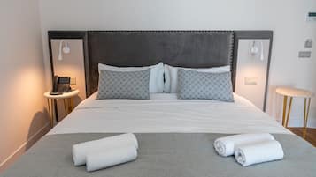 Una cama bien hecha con cabecera gris, dos mesitas de noche con lámparas y una pequeña mesa redonda con lámpara.