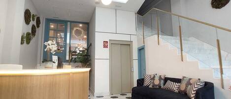 Een moderne receptie met een houten balie, een zwart-wit betegelde vloer met honingraatpatroon, een zwarte bank met decoratieve kussens en een trap met glazen leuning.