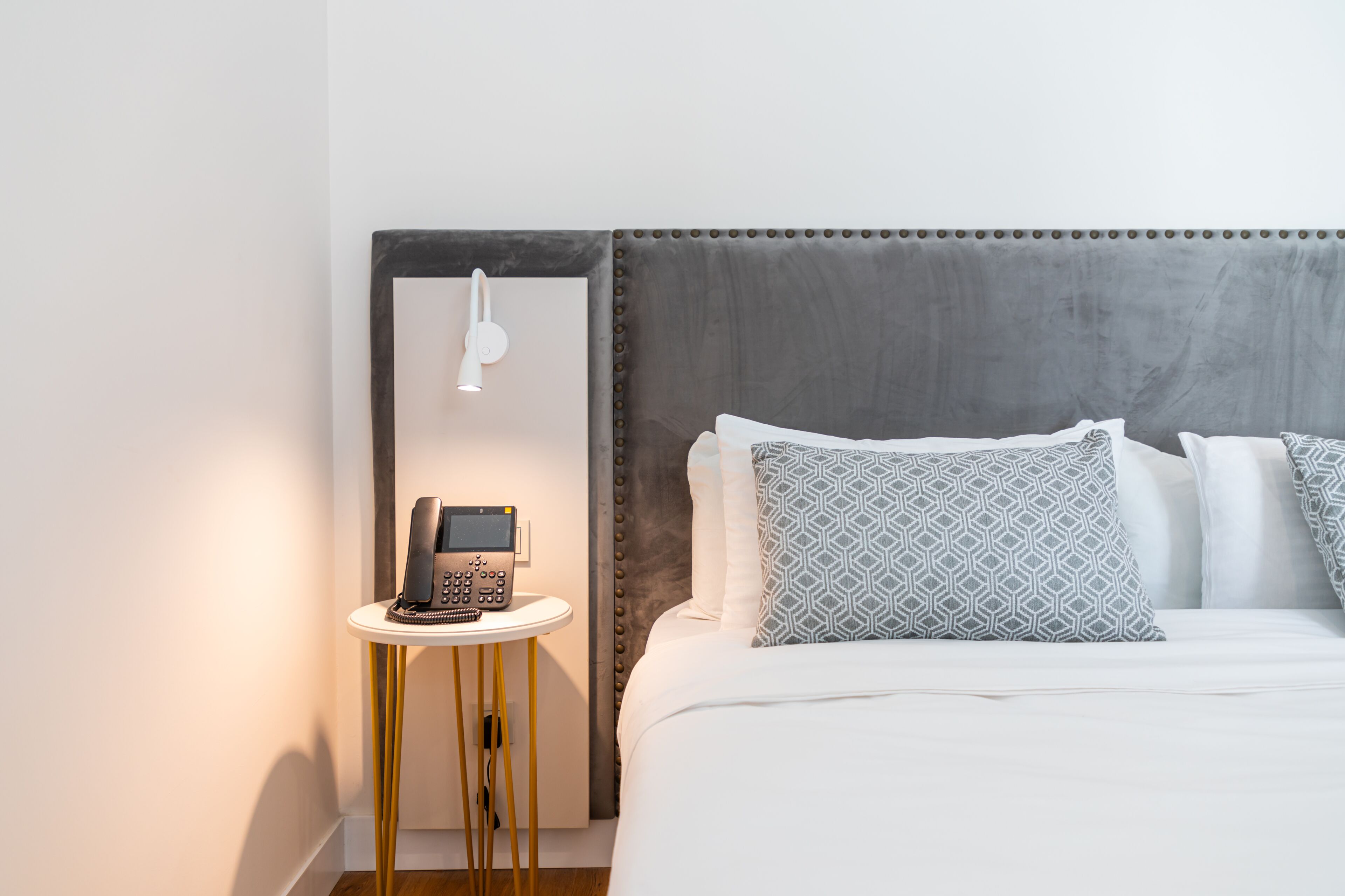 A hotel room with a bed, a bedside table with a phone, and a wall-mounted light.