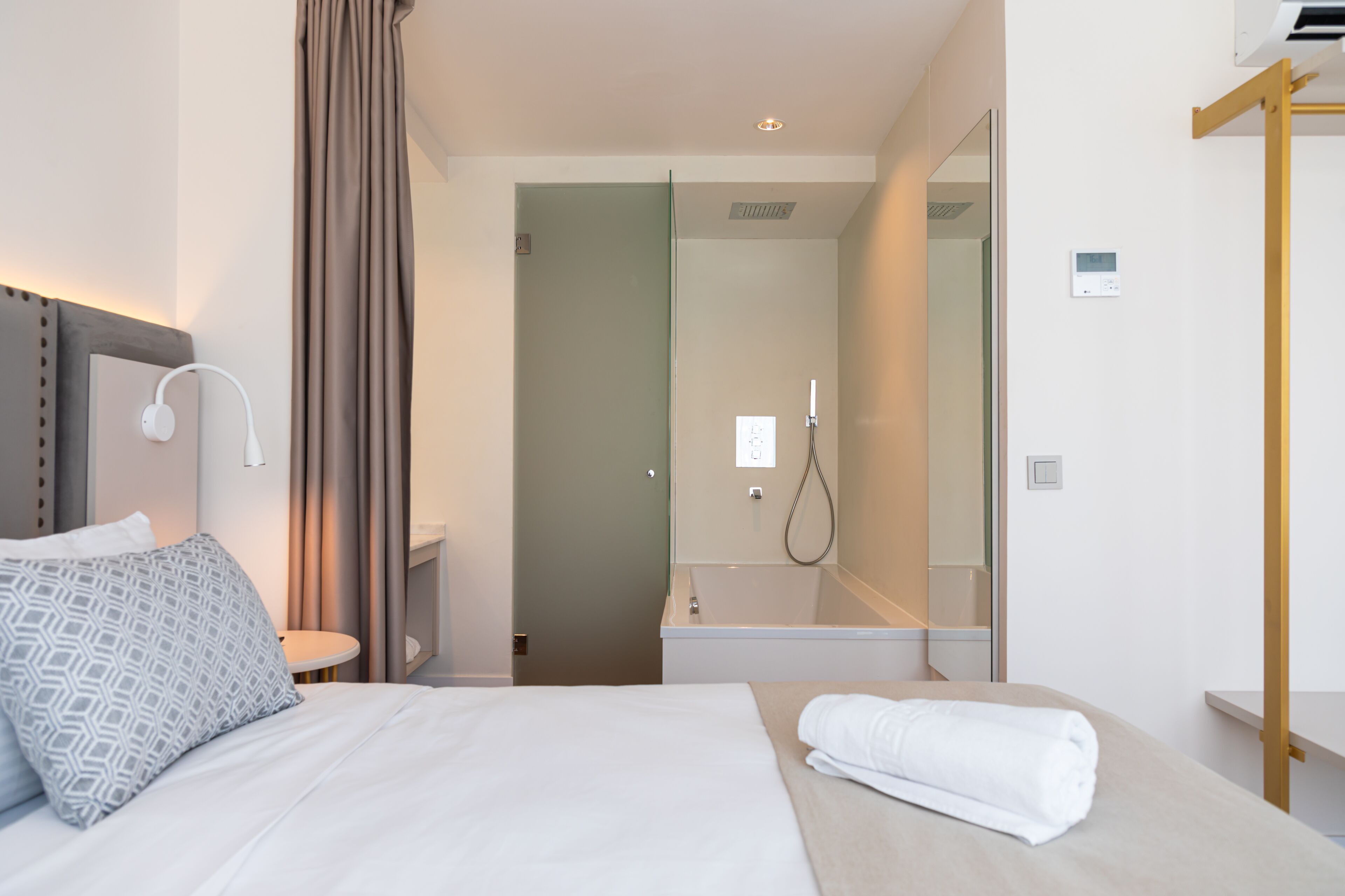 Habitación de hotel con cama, mesita de noche, lámpara, toalla, zona de ducha y espejo.