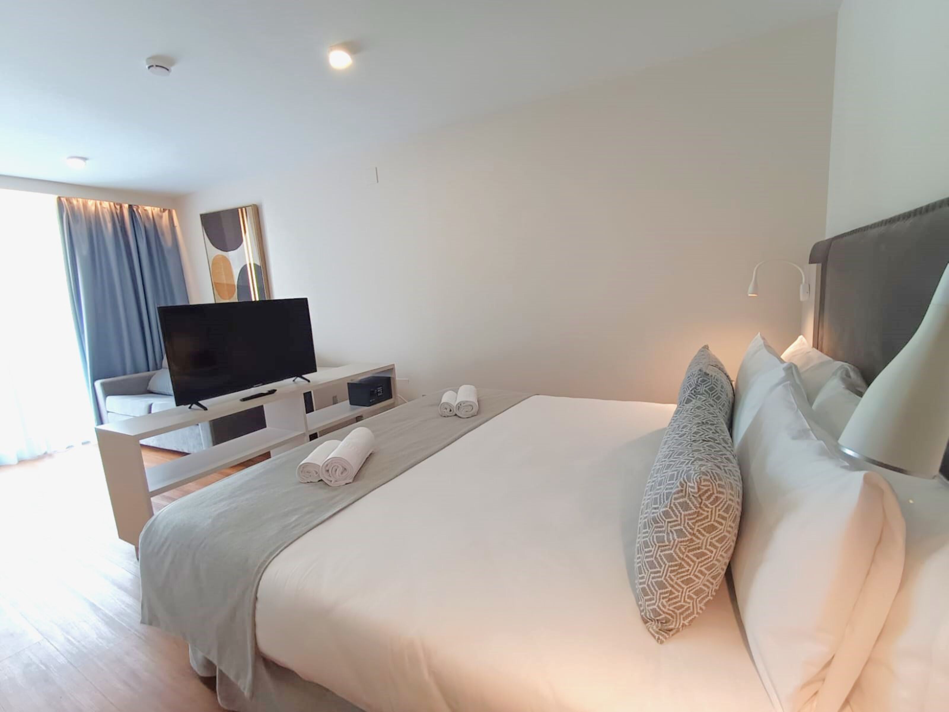 Habitación de hotel moderna con una cama grande, un televisor de pantalla plana, una mesita de noche con lámpara y vistas al exterior.