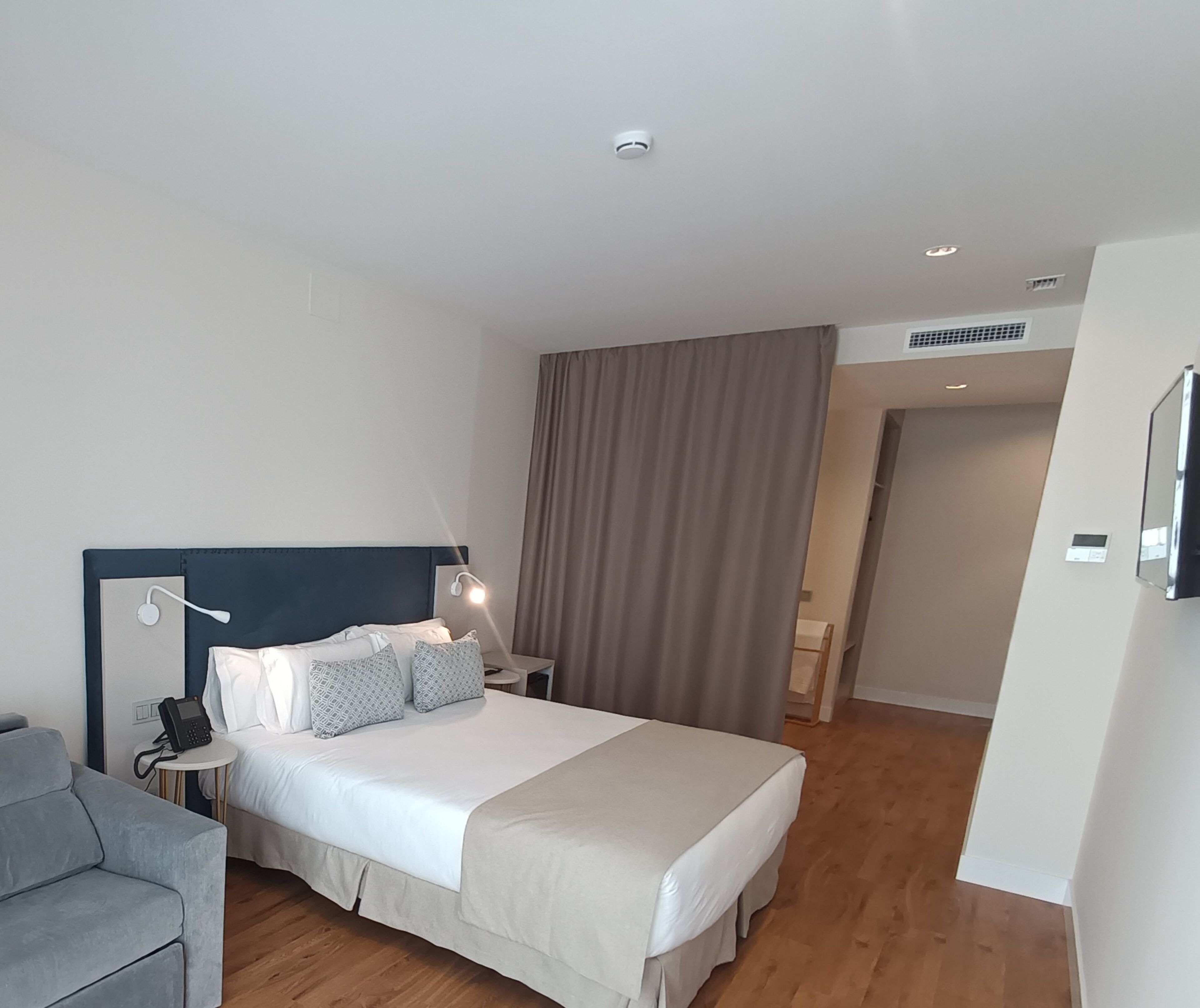 A modern hotel room with a neatly made bed, a bedside table with a lamp, a phone, and a grey sofa.
