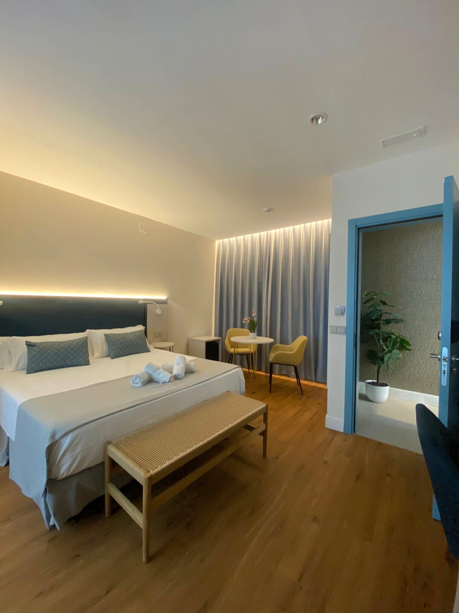 Una habitación de hotel moderna con una cama grande, mesitas de noche, una pequeña mesa redonda con sillas, un banco y una planta en maceta.