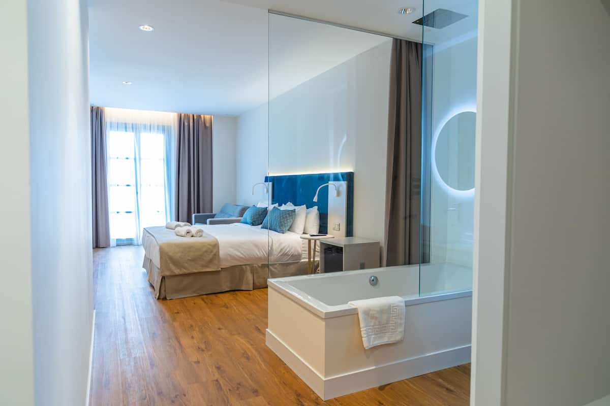 Una habitación de hotel moderna con una cama grande, una bañera con mampara de vidrio y un espejo amplio.