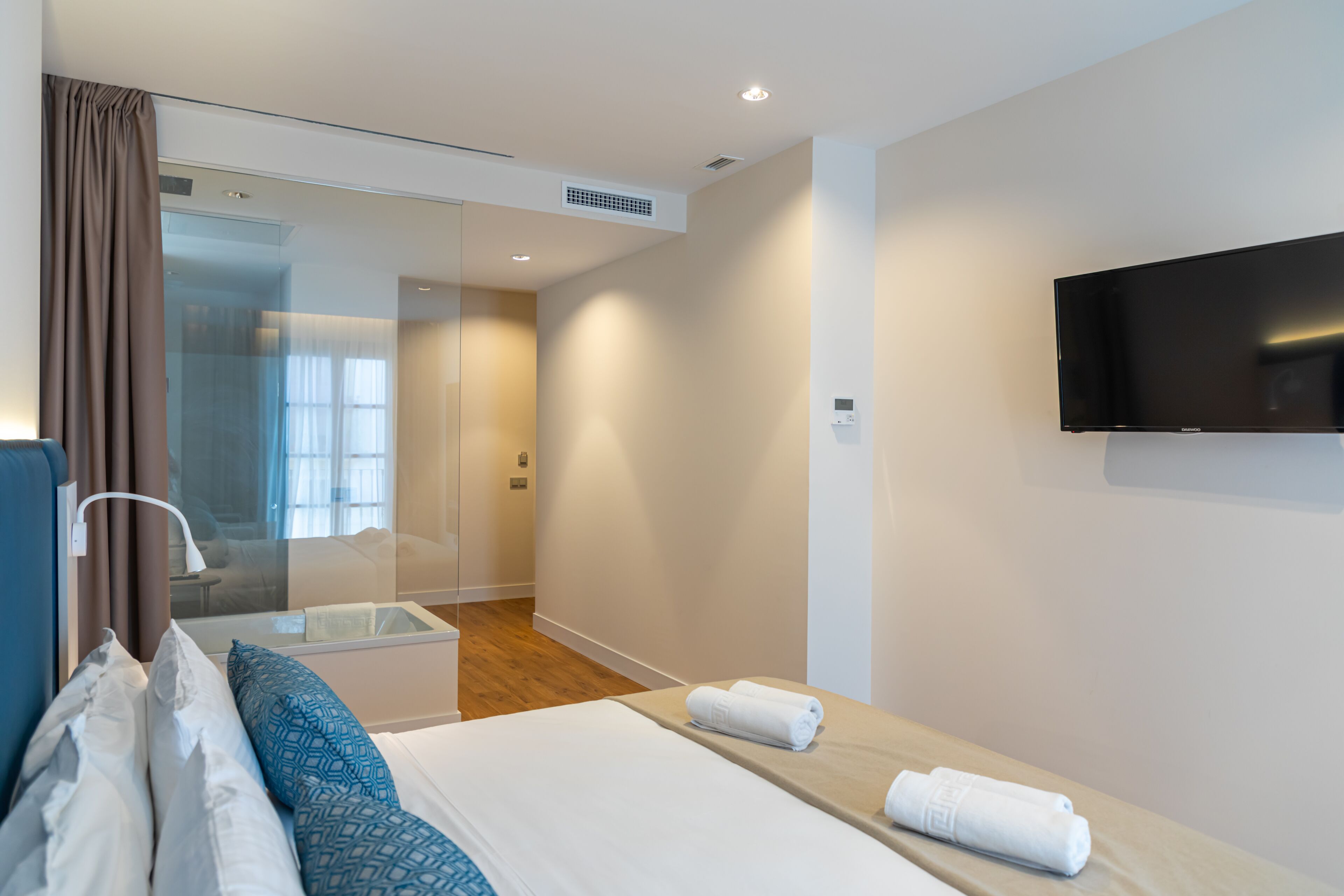 Ein modernes Hotelzimmer mit einem großen Bett, einem an der Wand montierten Flachbildfernseher, einer Duschkabine aus Glas und einem Holzboden.