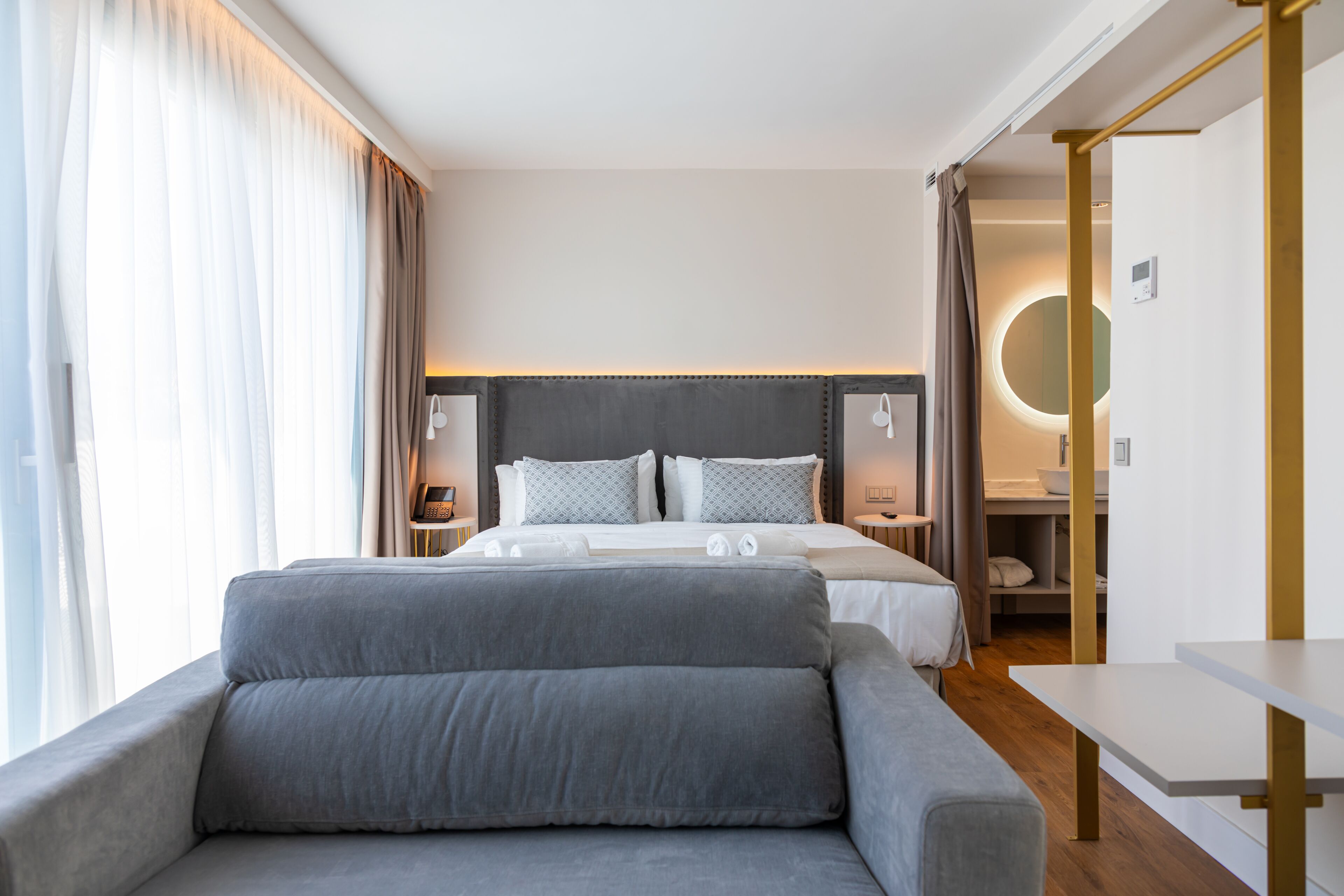 A modern hotel room with a large bed, a sofa, a bedside table, and a view of the city.