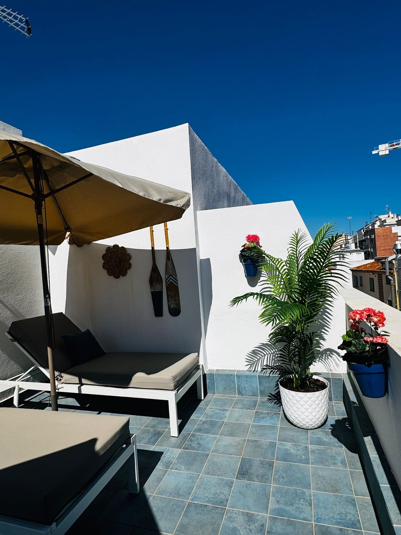 A rooftop terrace with a sun lounger, a table, a hanging chair, a potted plant, and a wall-mounted shelf with flowers.