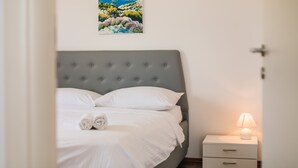 A neatly made bed with white linens and a grey headboard, a white bedside table with a lamp, and a painting of a coastal landscape on the wall.