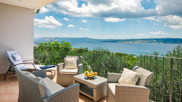 A balcony with wicker furniture, a table with fruit, and a view of the sea and mountains.