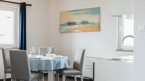 A dining area with a table set for two, chairs, a painting on the wall, and a window with curtains.