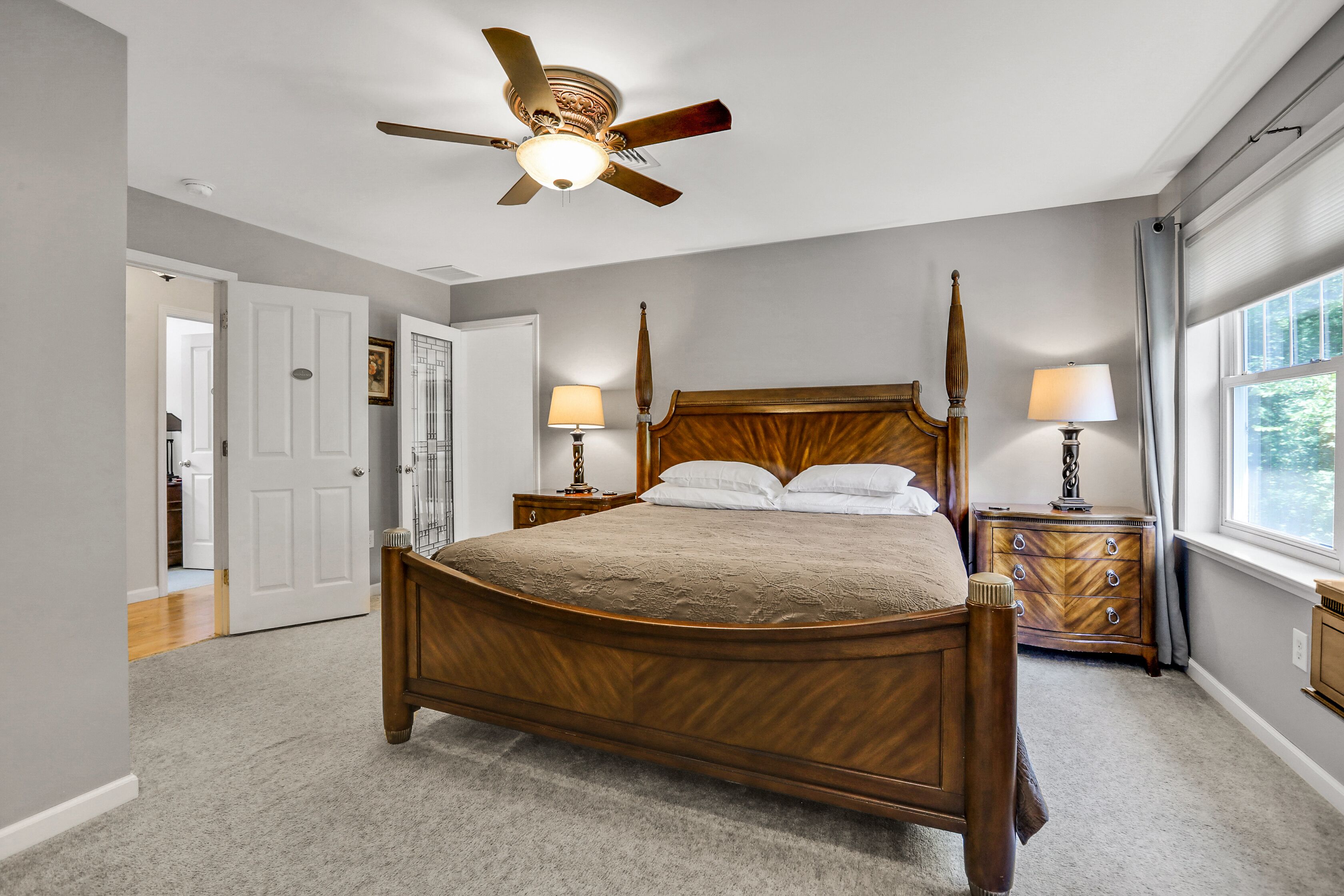 A four-poster bed with a wooden headboard, two bedside tables with lamps, a ceiling fan, and a window with curtains.