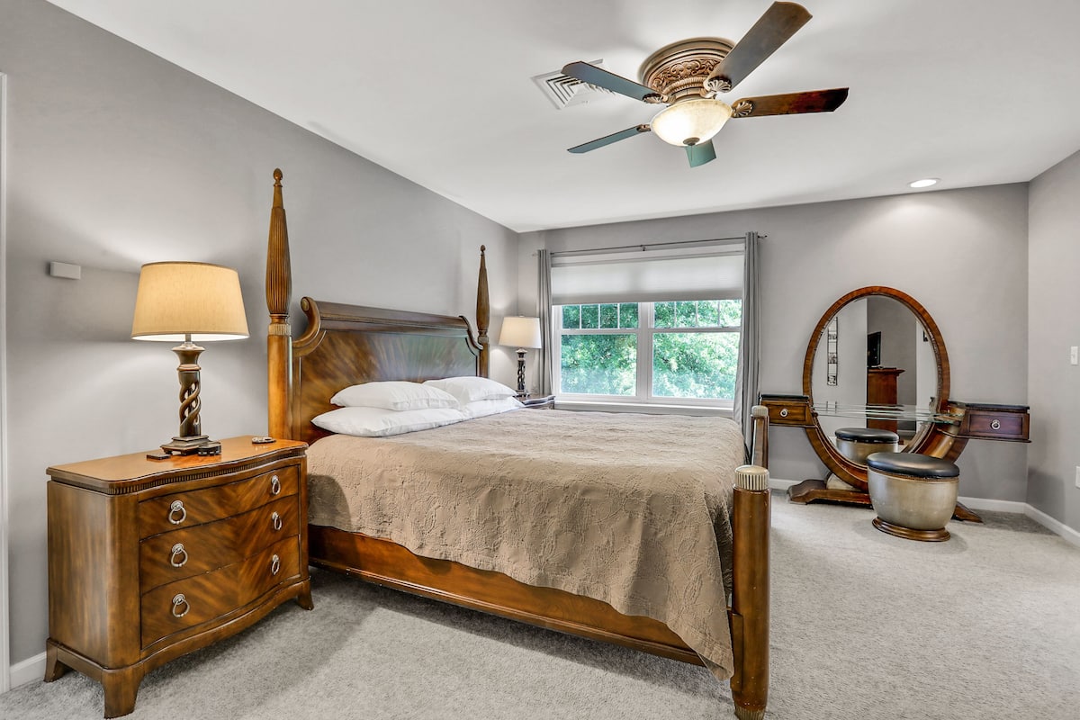 A bedroom with a wooden bed, a nightstand with a lamp, a ceiling fan, a large window, and a vanity with a mirror.