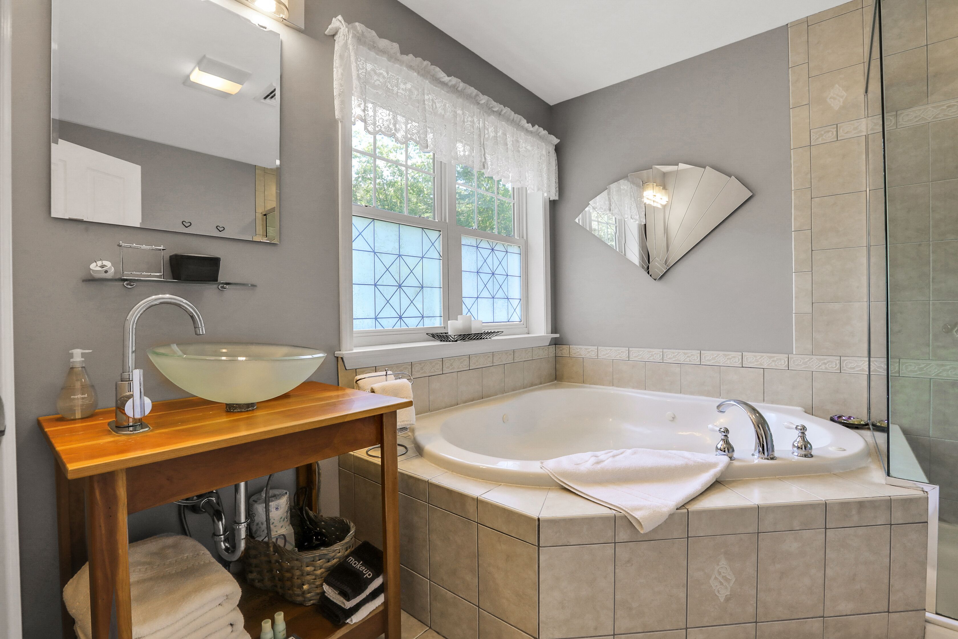 Ein Badezimmer mit einer großen Badewanne, einem Glaswaschbecken, einem Waschtisch mit Spiegel und einem Fenster mit Spitzenvorhängen.
