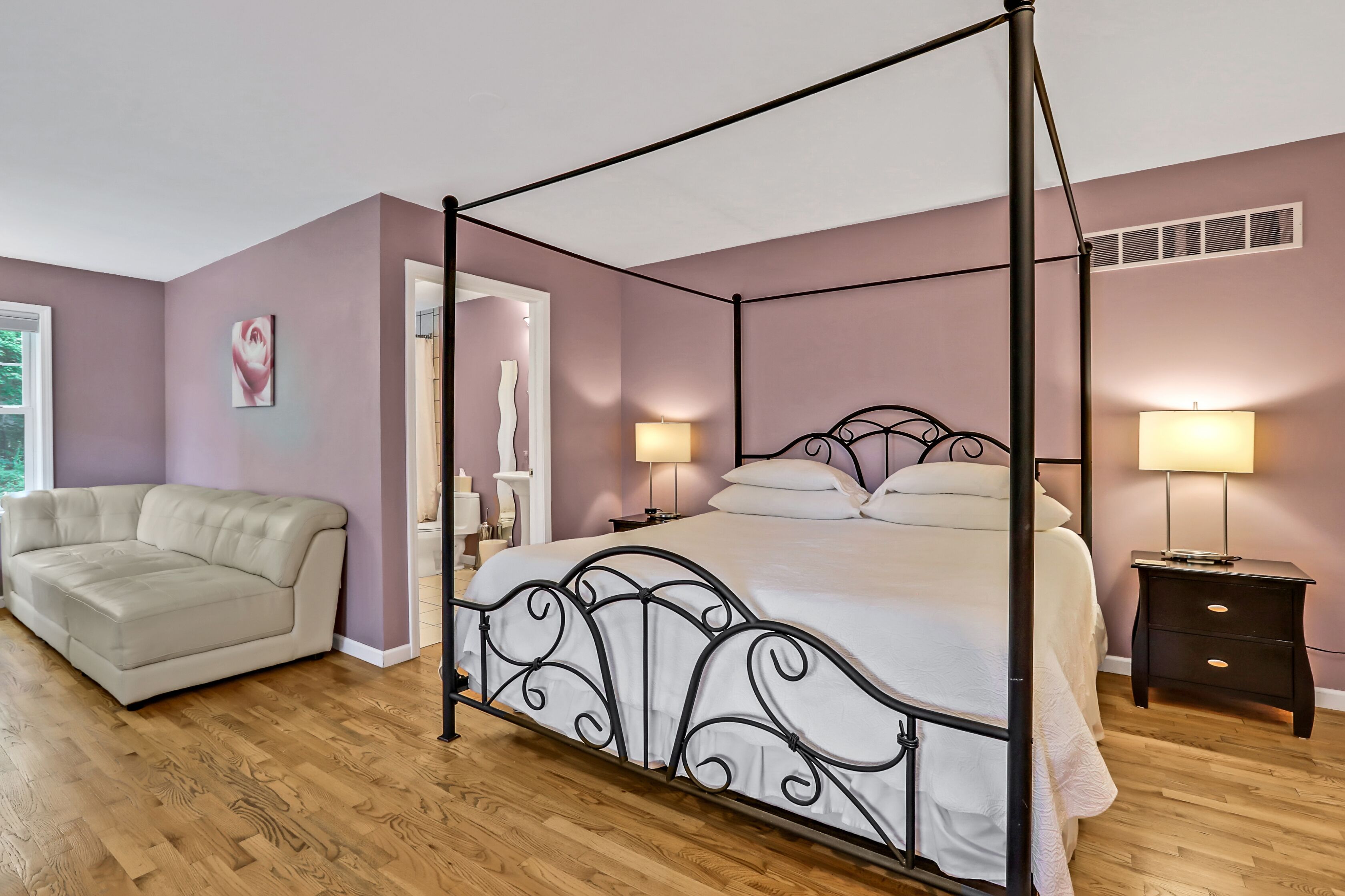 Uma cama com dossel, roupa de cama branca, duas luminárias de cabeceira, um sofá bege e piso de madeira.