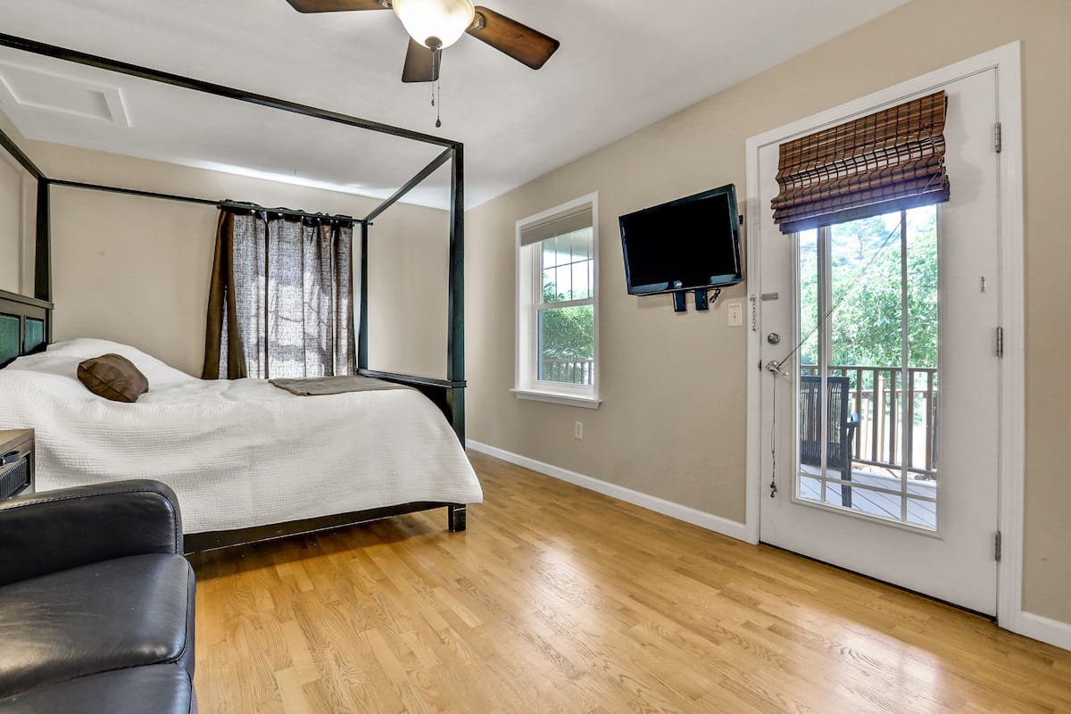 A bedroom with a four-poster bed, a ceiling fan, a television mounted on the wall, and a sliding glass door leading to an outdoor area.