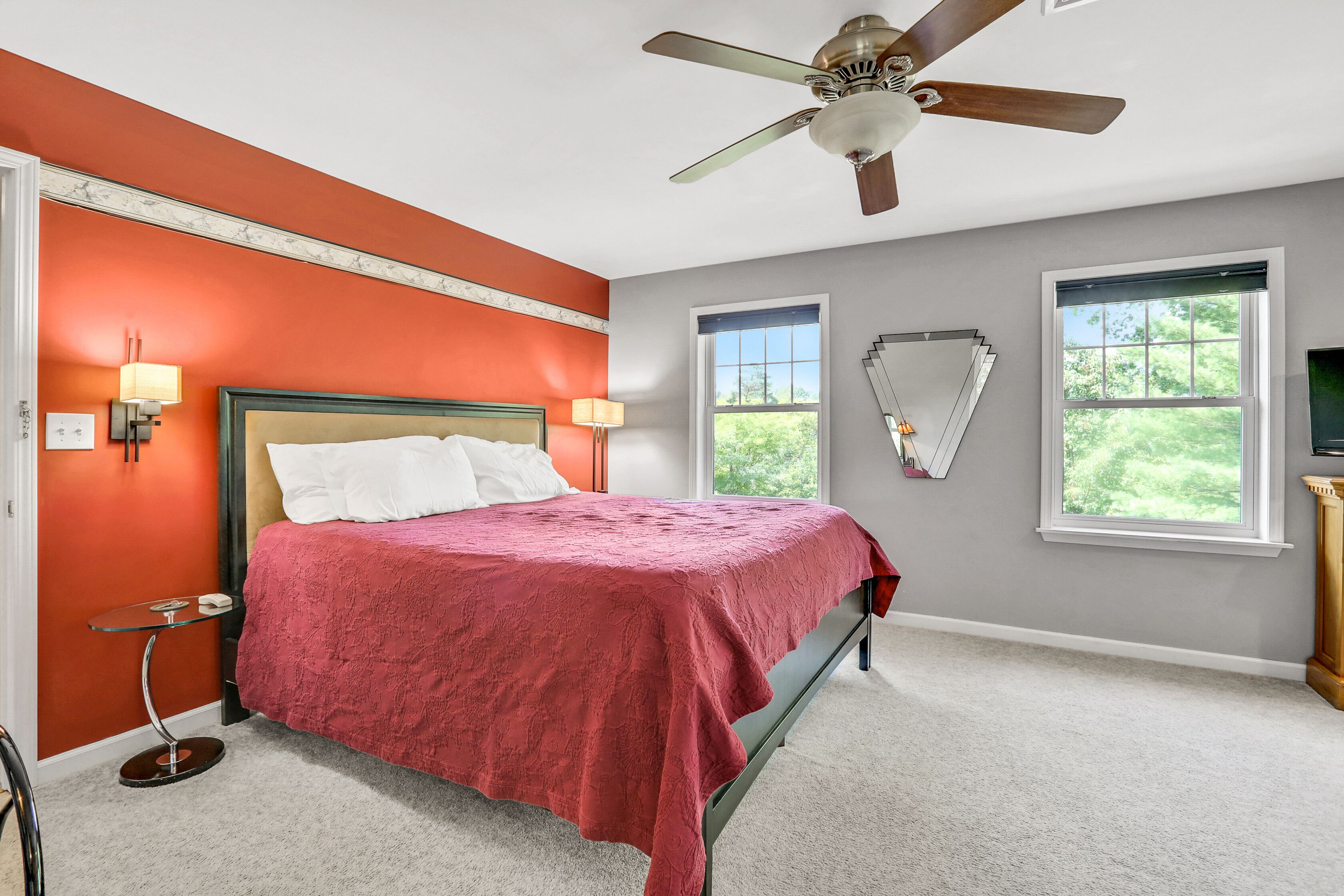 A bedroom with a bed, two windows, a ceiling fan, and a nightstand with a lamp.