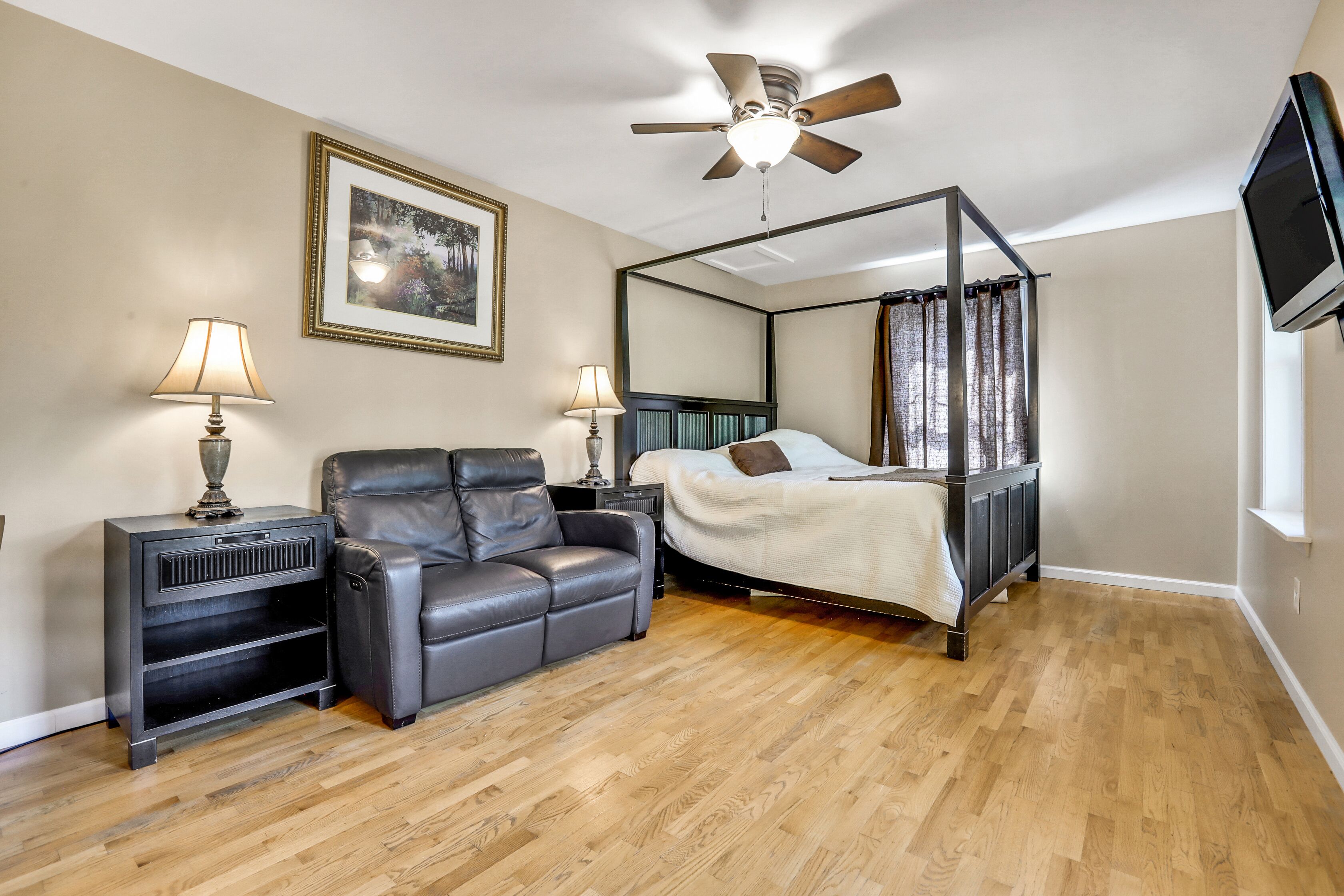 A bedroom with a four-poster bed, a leather sofa, two bedside tables with lamps, a ceiling fan, and a TV mounted on the wall.