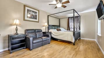 A bedroom with a four-poster bed, a leather sofa, two bedside tables with lamps, a ceiling fan, and a TV mounted on the wall.