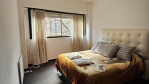 A bedroom with a bed, a window with curtains, a radiator, and a ceiling light. - Exquisito 3 ambientes Congreso (Buenos Aires)