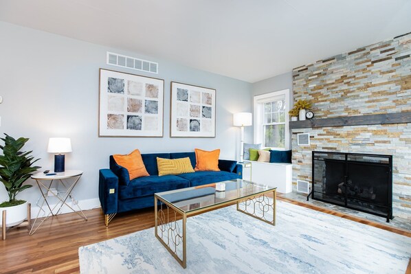 A living room with a blue sofa, a glass coffee table, a fireplace, and framed artwork on the wall. - Backyard Vibes-Walk to ND-Dogs Welcome-BBall Hoop-Fire Pit-Sleeps 12-2024 Reno (South Bend)