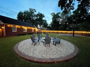 A backyard with a circular seating area, a basketball hoop, and a house with string lights. - Backyard Vibes-Walk to ND-Dogs Welcome-BBall Hoop-Fire Pit-Sleeps 12-2024 Reno (South Bend)