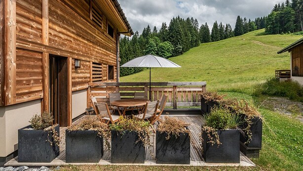 Een houten chalet met een terras, inclusief een tafel en stoelen, omgeven door plantenbakken met beplanting.