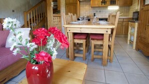 Une table de salle à manger en bois avec des chaises, un vase rempli de fleurs et un coin cuisine avec des meubles en bois et un dosseret carrelé.