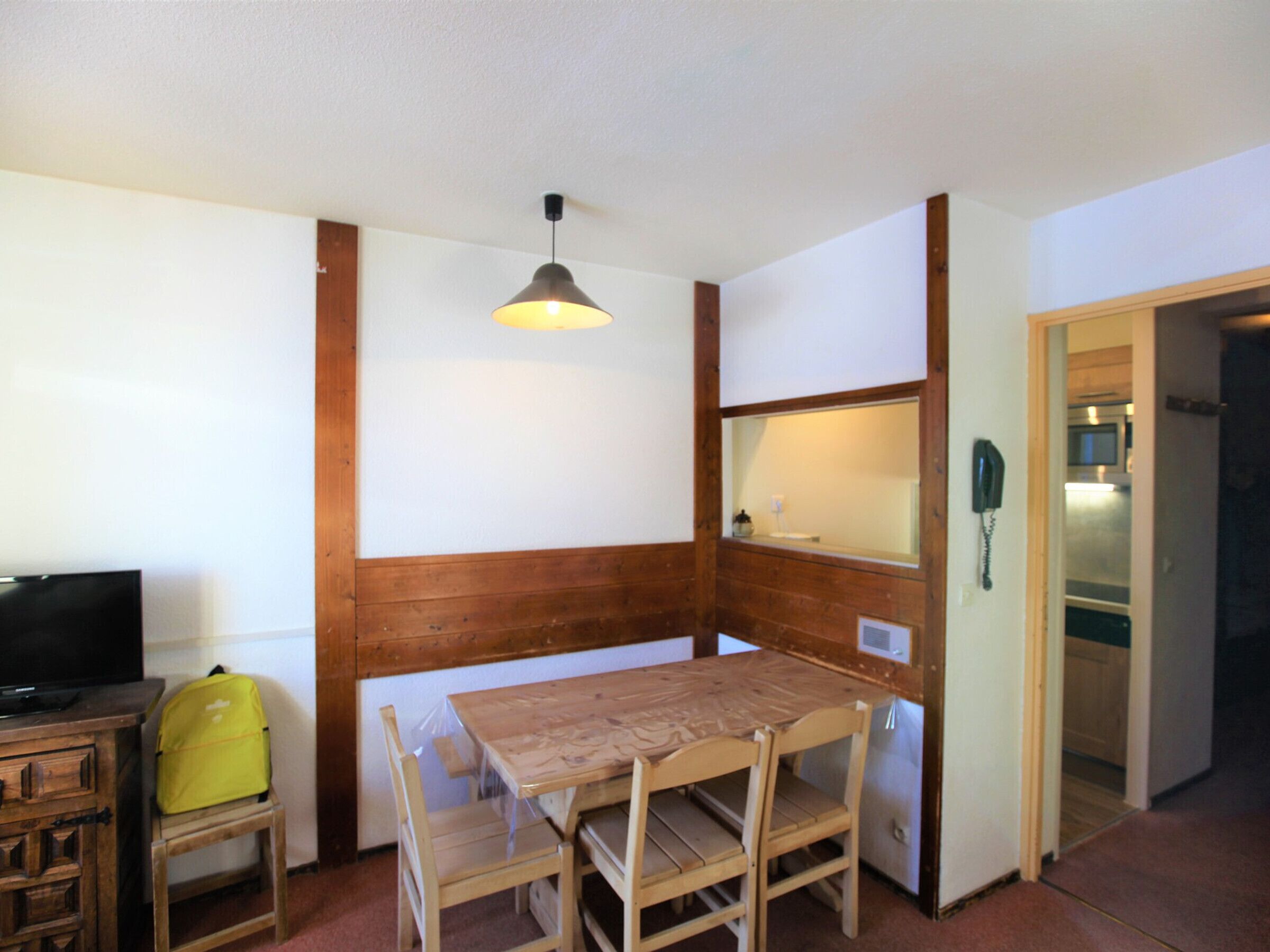 Ein gemütlicher Raum mit einem Holztisch und Stühlen, einem Fernseher auf einer Holzkommode, einer Hängeleuchte und einem Telefon an der Wand.